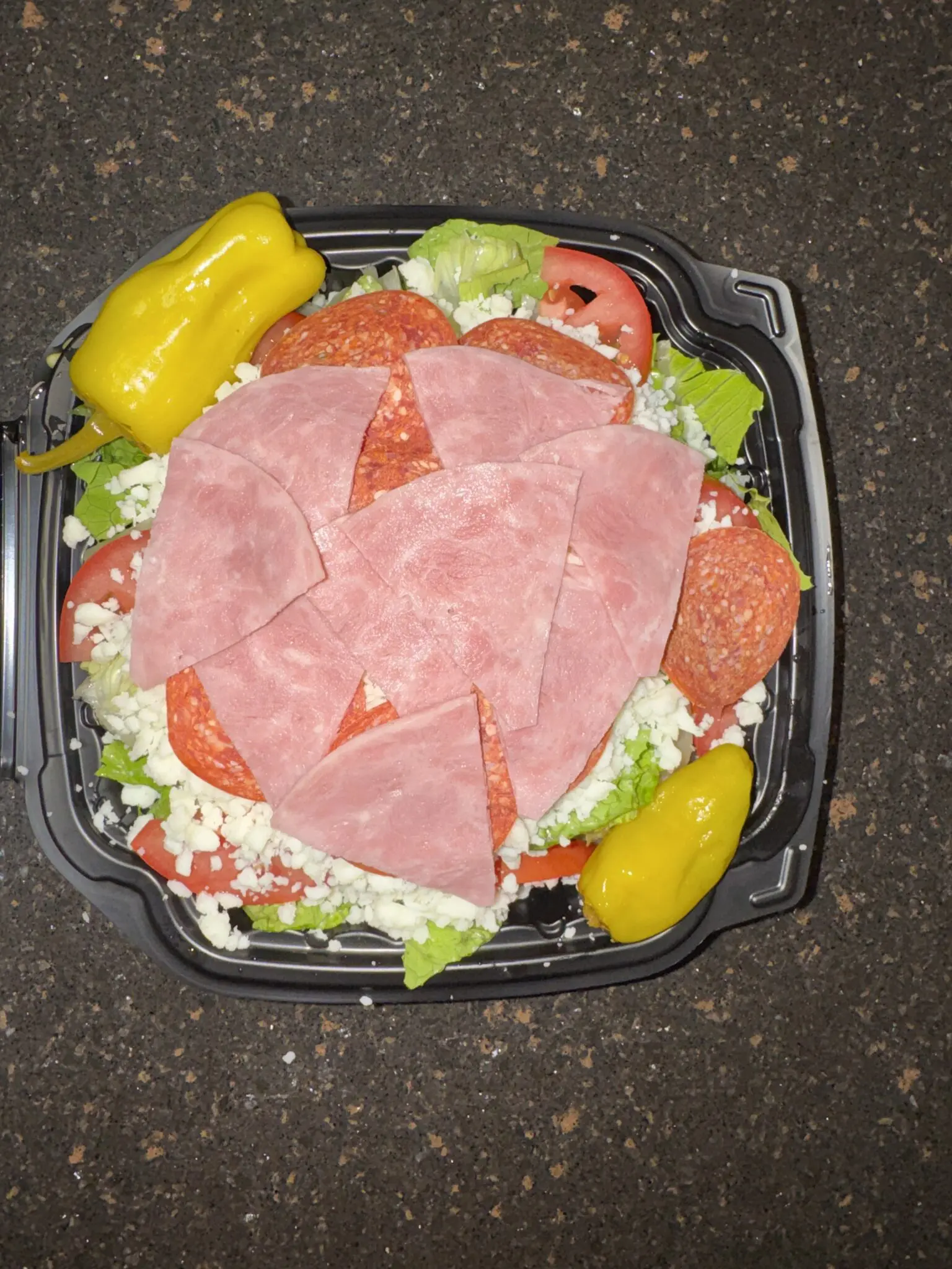 Fresh salad topped with sliced deli meats and pepperoncini peppers in a black container.