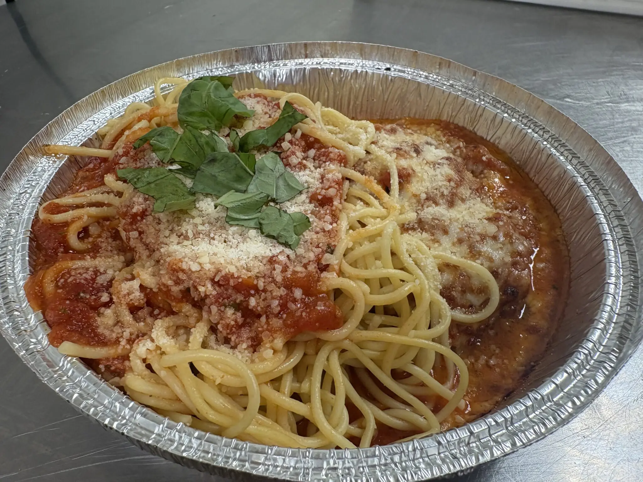 Spaghetti topped with tomato sauce, grated cheese, and fresh parsley in a foil container.