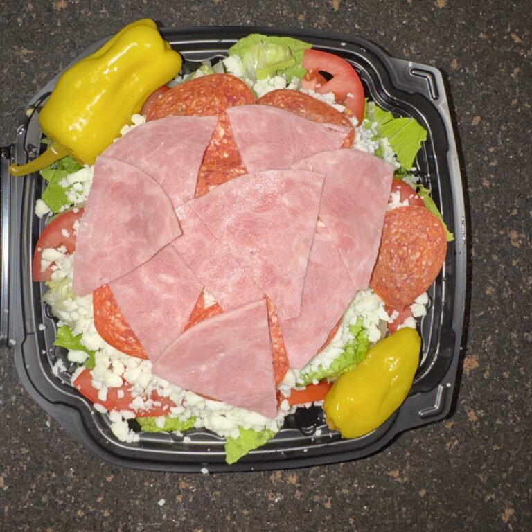 Fresh salad topped with sliced deli meats and pepperoncini peppers in a black container.